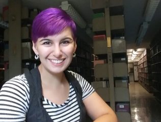 Yaine Neyhard sits in the library, wearing a striped shirt, with short, purple hair.