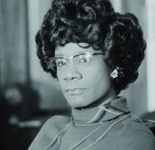 A black and white photo of Shirley Chisholm in 3/4 profile from the shoulders up. 
