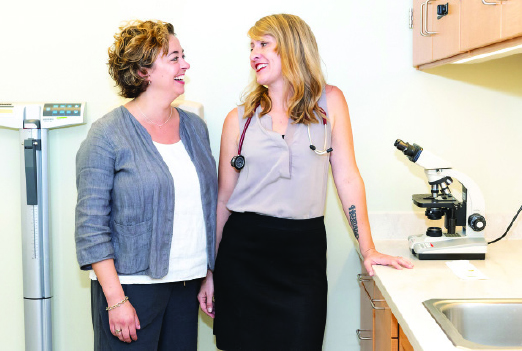 Katharine Smith ’05 and Becky Stephens ’03 look at each other, smiling. They stand against a white wall in their office between a digital scale and a microscope sitting on the counter next to a sink.