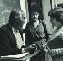 Chisholm stands in front a chalkboard, smiling while talking with a student, while another student looks on.